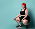 Overweight ginger model in red headband, black top, shorts, sneakers. Squatting posing sideways against blue studio background