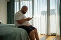 Overweight guy hanging in smartphone on resort sitting in bed in hotel room on vacation.