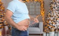 Overweight man measuring his waist in room with Christmas trees after holidays, closeup Royalty Free Stock Photo