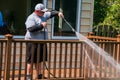 Overweight man wearing white baseball cap power washing the railing of a brown wooden deck in a backyard.