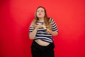 Overweight plump satisfied joyful woman with naked belly and close eyes in striped clothes eating potato chip. Addiction