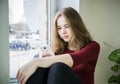 Overweight teenager girl with long blond hair is sitting by the window on the windowsill with a phone in her hand. The concept of