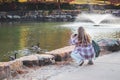 Overweight woman with long streaked hair crouches down by pond with fountains on fall day to take phone picture of geese in water