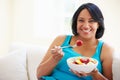 Overweight Woman Sitting On Sofa Eating Bowl Of Fresh Fruit