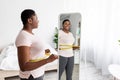 Overweight young black woman measuring waist with tape near mirror at home, satisfied with slimming result, free space Royalty Free Stock Photo