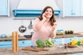 overweight young woman with wooden spoon in hand talking on smartphone while standing at table with fresh vegetables in kitchen Royalty Free Stock Photo