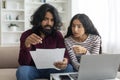 Overwhelmed couple sitting together on couch, using laptop, holding papers