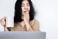 Overworked Asian businesswoman yawning at office desk hand holding a cup of coffee feeling tired and sleepy