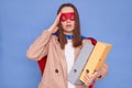 Overworked sad tired brown haired woman wearing superhero costume holding documents in folders posing isolated over blue