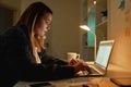 Overworking. Side view of young caucasian woman working from home in dark late night, using laptop and drinking coffee