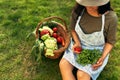 Owner female gardener with a basket with freshly picked ecological vegetables sitting on the grass at the farm. A young farmer