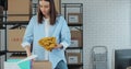 The owner of an online store works on a computer at her desk in a warehouse. An employee packs a yellow jumper in a room