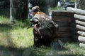 Paintball game in the forest. Summer. A girl in camouflage clothes with a weapon in her hands is looking out from behind a wooden