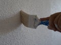 Close-up of blue paintbrush painting a white wall. The wall has popcorn ceiling texture. Man holding the tool has his hands Royalty Free Stock Photo