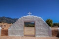 Painted doorway topped by cross with mountains and blue sky and man carrying bucket viewed through it at Taos Pueblo in New Mexico