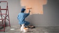 Painter with cap squatting painting a wall with paint roller, red ladder and buckets near. repair, building and home Royalty Free Stock Photo