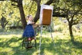 Painting on canvas using easel in the green summer garden with bright sunlight. Caucasian little artist girl, wearing Royalty Free Stock Photo