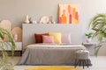 Painting on grey headboard of bed with colorful cushions in bedroom interior with stool. Real photo