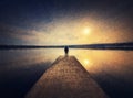 Painting of a man standing alone on the pier facing the sunset reflected on the calm lake water. Scenic view with a lonely person Royalty Free Stock Photo