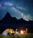 Pair backpackers sitting near campfire and looking to the camera under incredibly beautiful starry sky at night camping