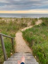 A pair of bare feet, crossed ankles pointing down a set of weathered steps to a strip of sandy path which leads to the ocean. Royalty Free Stock Photo