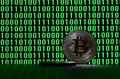 A pair of bitcoins lies on a cardboard surface on the background of a monitor depicting a binary code of bright green zeros and on