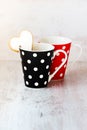 A pair of black and red polka dotted coffee cups with a heart shaped homemade cookie on the edge over white wood background.