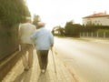 A pair of elderly people walk along the sidewalk along the road holding hands. Grandfather and grandmother on a walk in a