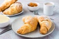 A pair of fresh crispy croissants on a plate, a cup of coffee and bowls of jam and butter on a light textured background. Coffee