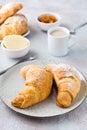 A pair of fresh crispy croissants on a plate, a cup of coffee and bowls of jam and butter on a light textured background. Coffee