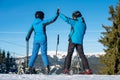 Pair giving high five to each other, smiling, standing with skis on mountain top at a winter resort with ski lifts