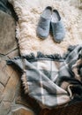 The pair of gray home slippers on the natural white sheep sheepskin with warm plaid dropped on the stone floor in the cozy bedroom