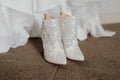 Pair of pristine white wedding shoes on a brown carpet floor with a wedding dress in the background