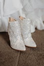 Pair of pristine white wedding shoes on a brown carpet floor with a wedding dress in the background