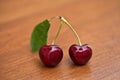 Two red ripe cherries with stems and a leaf on a wood table