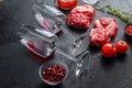 Pair red wine glass near raw organic beef steak cuts, with rosemary, spicy chili oil on black textured background side view