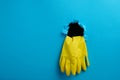 A pair of yellow latex house cleaning gloves sticking out of the torn hole of the blue paper background