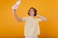 Pale girl with arm tattoo taking picture of herself in yellow room. Studio portrait of seious curly lady making selfie..