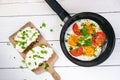 Pan of fried eggs with tomatoes, cheese, spring onion, herbs on a white table. Bread with spread. White wooden table. Concept of f Royalty Free Stock Photo