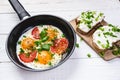 Pan of fried eggs with tomatoes, cheese, spring onion, herbs on a white table. Bread with spread. White wooden table. Concept of f Royalty Free Stock Photo