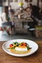 Pan seared Cod fish fillet with tomato, bean and carrot in gravy sauce. Served in white plate on wooden table with blur people.