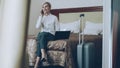 PAN of smiling businesswoman in white shirt using on laptop and talking at mobile phone while sitting on bed in hotel Royalty Free Stock Photo