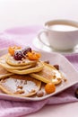 Pancakes with raspberries, physalis, hazelnut and honey on pink plate, sprinkled with powdered sugar, with fork and cup of coffee