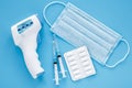 Pandemic of coronavirus COVID-19. Protective surgical mask, pills, electronic infrared thermometer and two syringe on blue