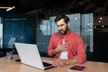 Panic attack in the workplace. A young man sits in the office at the table, works on a laptop, holds his hand to his