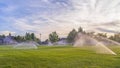 Pano frame Sprinklers watering green grassy field with homes and cloudy blue sky background