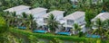 Panorama aerial riverside white painted tropical villas, swimming pool, lounge chairs, lush green coconut palm trees, row upscale