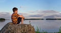 Panorama of a boy sitting on rock in summer evening sunlight