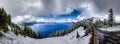 Panorama of Crater Lake with a road on the right, deep blue water, snow covered pine trees and dramatic clouds