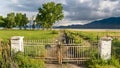 Panorama Dirt road with rusty metal gate on a vast grassy field in front of a lake